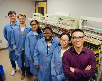 Five lab members and Feng Lin smile in the lab. The lab members wear blue lab coats and safety glasses, and Lin wears a red shirt. 