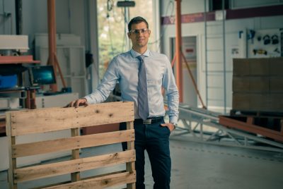 Laszlo Horvath stands in a large, equipment-filled room holding up a wooden pallet.