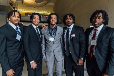 Five Black male students in suits and ties pose together in the atrium of Goodwin Hall.