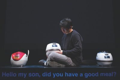 South Korean theatremaker Jaha Koo, an Asian man with short dark brown hair and round glasses, wears a black shirt and sits on top of a table between two Cuckoo brand rice cookers, another in his lap. Projected subtitles read “Hello my son, did you have a good meal?”