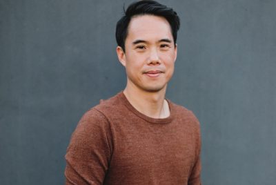 Author Charles Yu, a young Asian man with short dark hair, wears a rust brown T-shirt and smiles towards the camera in front of a medium grey background.