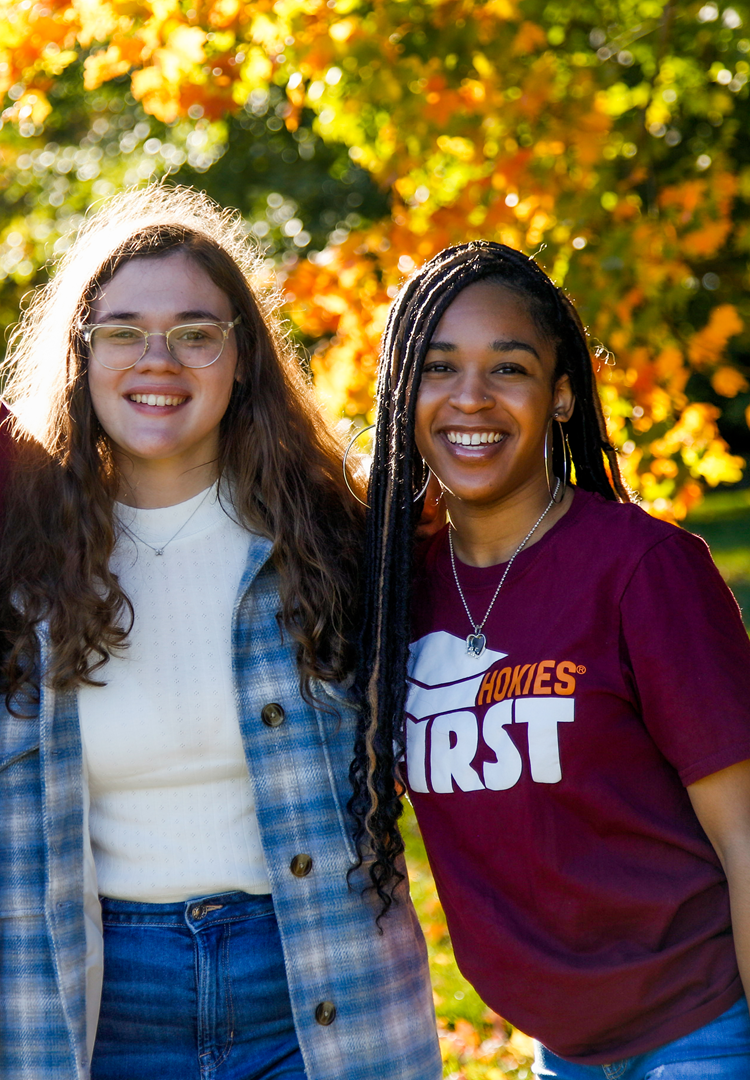 Two students pose together in front of autumn foliage at Virginia Tech. Two students stand side-by-side, arms around each other's back, with orange, red, and green foliage behind them.